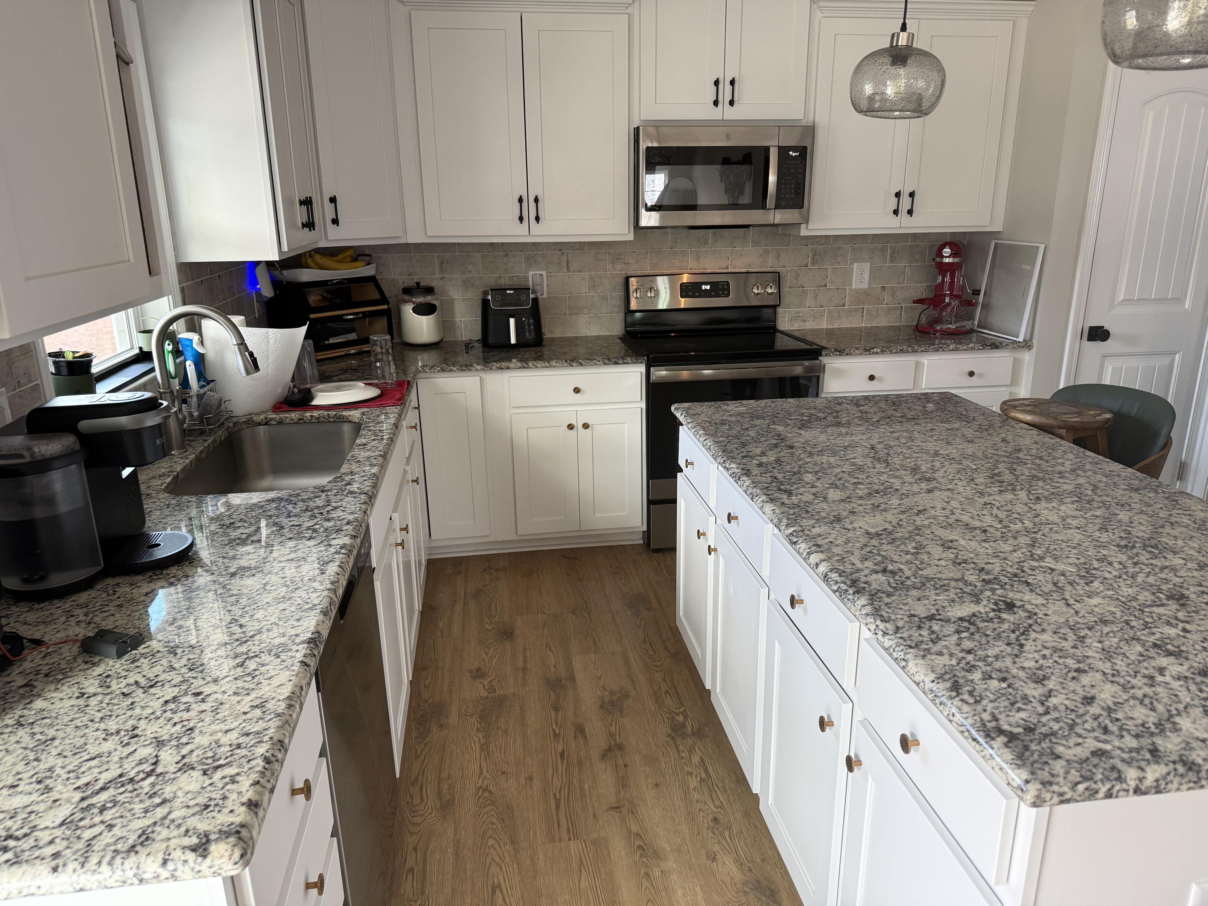 A bright residential kitchen features white shaker cabinets, speckled granite countertops, and a large central island with pendant lighting.