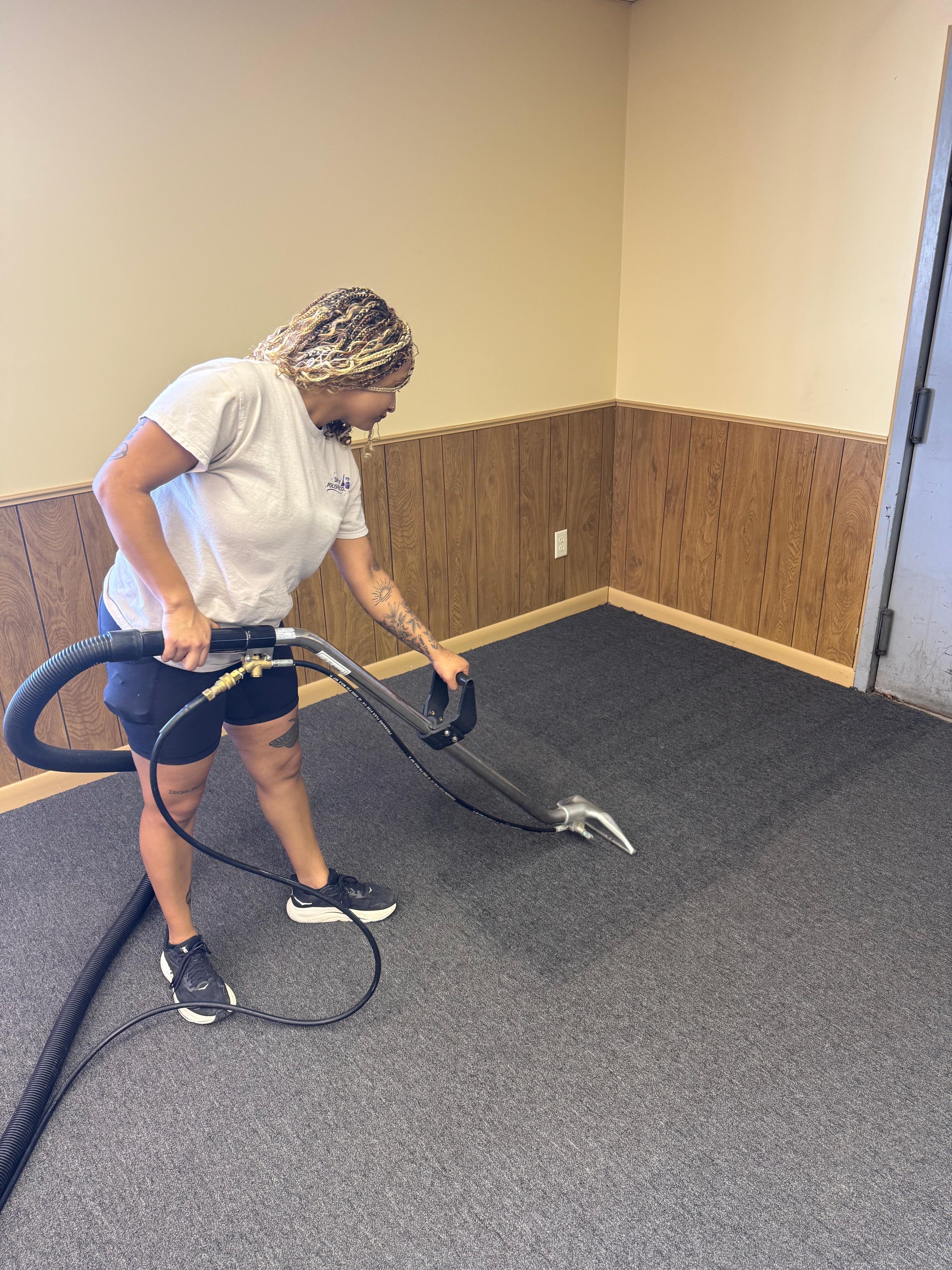 A technician with blonde braids operates a carpet cleaning wand on a section of dark grey carpeting next to light wood flooring.